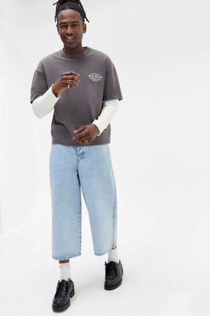 Mand med dreadlocks iført lagdelte grå langærmede og kortærmede skjorter, lyseblå vide jeans, hvide sokker og sorte sko på hvid baggrund.