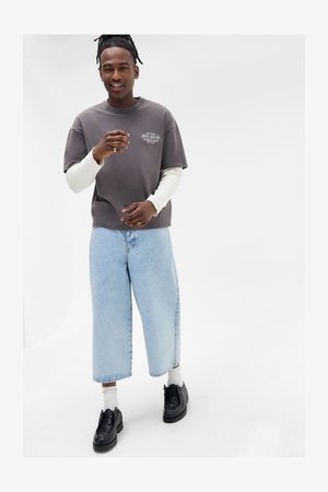Mann med dreadlocks iført lagdelte grå langermede og kortermede skjorter, lyseblå vide jeans, hvite sokker og svarte sko på hvit bakgrunn.