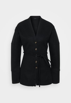 Melna jaka ar apvalku, izgatavota no mīksta auduma; tai ir dziļa V-veida kakla izgriezums, lielas piedurknes, seši pogas priekšpusē un atbilstoša sasiešana viduklī.