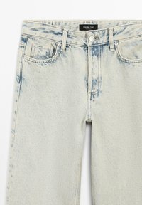 Světle modré džíny z denimu s vybledlým povrchem, obsahující pět kapes, zipové zapínání a knoflíkový uzávěr. Materiál má mírnou texturu.