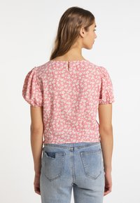 Lyserød bluse med blomsterprint og pufærmer, der har en nøglehulslukning på ryggen. Sammen med lyseblå denimjeans, der viser baglommer.