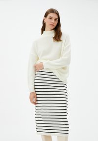 Kerman neulepusero korkealla kauluksella, yhdistetty valkoiseen hameeseen, jossa on ohuita mustia vaakaraitoja. Minimalistinen muotoilu, rento istuvuus.