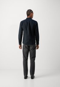 Camisa de manga larga azul oscuro con un cuello estándar, puños abotonados y jeans grises de corte recto. Modelo de pie con la espalda hacia nosotros.