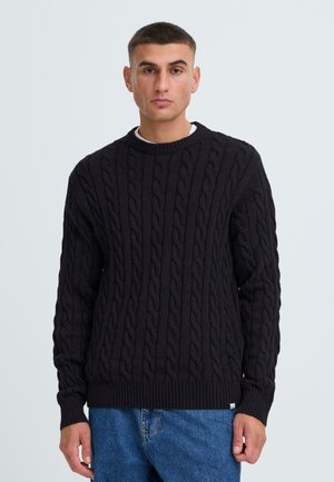 Schwarzer Strickpullover mit Rundhalsausschnitt, der über eine strukturierte Musterung und gerippte Bündchen sowie Saum verfügt. Getragen über hellblauen Jeans.