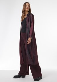 Camisa larga burdeos con un estampado texturizado, abotonada en la parte frontal, combinada con pantalones de pierna ancha burdeos a juego y botas negras gruesas.