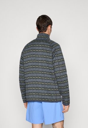 Mann mit dem Rücken zur Kamera, trägt eine gemusterte dunkelgrüne und graue Jacke und hellblaue Shorts vor einfarbigem Hintergrund.