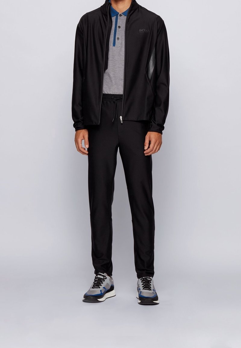 Homme portant une veste zippée noire, un pantalon noir à cordon, un polo gris avec un col bleu et des baskets grises avec des touches bleues.