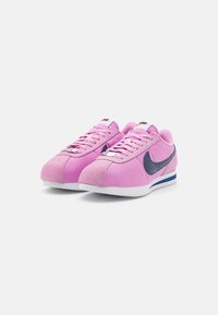 Rózsaszín sportcipő, velúr és szintetikus felsőrészből, tengerészkék swoosh-al, fehér talppal és textúrált fűzőkkel. Alacsony profilú, kerek orrú kialakítás.