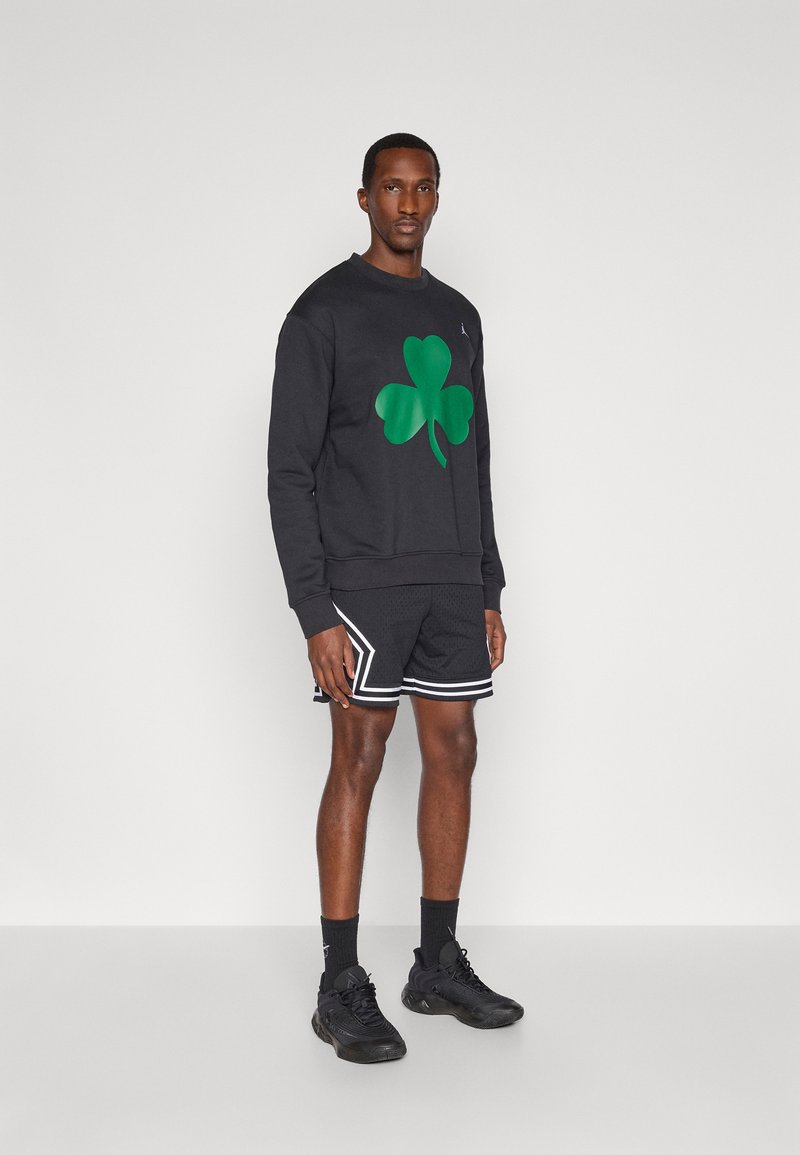 Man standing wearing black sweatshirt with large green shamrock, black athletic shorts with white trim, black socks, and black sneakers against white background.
