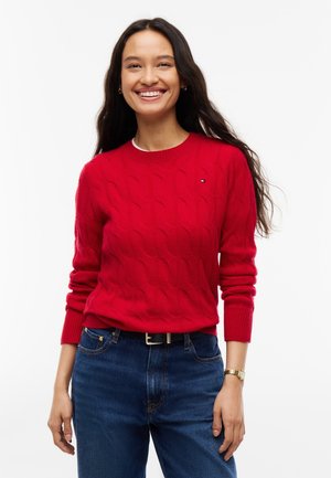 Mujer sonriente con el cabello largo y oscuro, vestida con un suéter rojo de punto trenzado, jeans azules, cinturón negro y un reloj de pulsera dorado sobre fondo blanco.
