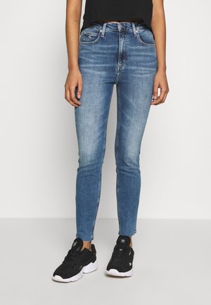 Frau trägt blaue Skinny-Jeans und schwarze Sneakers, steht vor einem schlichten hellen Hintergrund.