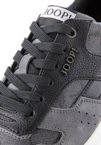 Gros plan sur une basket noire et grise avec des panneaux en cuir texturé et en tissu, des lacets noirs, et un logo de marque métallique "JOOP!" sur le côté.