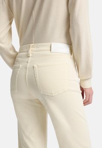 Fløjlsbukser i cremefarve med høj talje, baglommer og et hvidt mærke ved taljen. Tekstureret stof med lodrette riller.