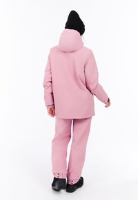 Roze waterdichte jas met een capuchon en elastische manchetten, gecombineerd met bijpassende broek. Voorzien van drukknoopsluitingen bij de enkels. Zwarte gebreide beanie gedragen.