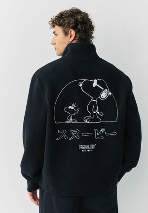 Schwarze Fleecejacke mit einem weißen Comic-Graphic von Snoopy und einem Vogel auf dem Rücken. Das Design enthält japanischen Text und Logo-Akzente.