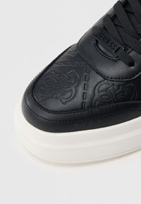 Mustast nahast spordijalats, millel on reljeefne logo, kontrastne valge tald, õmblustega detailid ja tekstuuriga viimistlus varba piirkonnas.