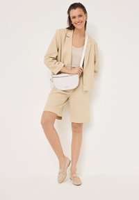 Beige linnen blazer met bijpassende shorts, gecombineerd met een licht tanktop. Witte getextureerde schoudertas en beige loafers met hardware accenten.