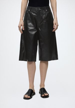 Person iført sorte knælange brede lædershorts og sorte åbne slippers, stående mod en hvid baggrund.