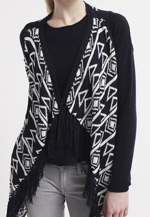 Cardigan aperto a strisce bianco e nero con frange, caratterizzato da design geometrici. Indossato sopra un maglione nero e jeans grigi.