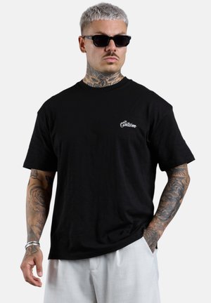 Mann mit kurzem platinblondem Haar, Sonnenbrille und Tattoos trägt ein schwarzes T-Shirt mit weißer Schrift und hellen Hosen, steht mit einer Hand in der Tasche.