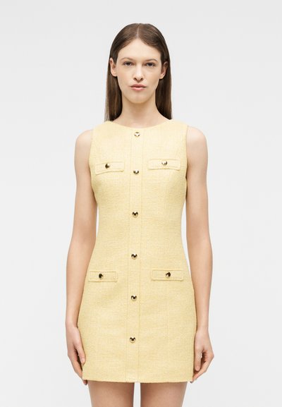 Νεαρή γυναίκα που φοράει αμάνικο, ανοιχτό κίτρινο φόρεμα με υφή, με χρυσά κουμπιά και τέσσερις μπροστινές τσέπες, στέκεται μπροστά σε φωτεινό φόντο.