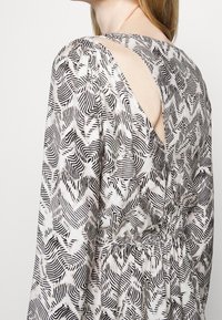 Černobílé vzorované šaty s dlouhými rukávy, úzkým pasem a detailem klíčové dírky na zádech. Tkanina vypadá lehce a texturovaně.