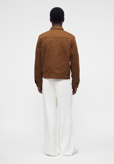 Homme debout de dos, portant une veste marron à motifs, un pantalon blanc large et des chaussures blanches sur un fond uni.
