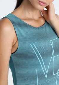 Un top senza maniche di colore acquamarina con un motivo a strisce orizzontali e un grande design grafico bianco. Realizzato in materiale liscio e elasticizzato.
