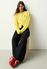 Keltainen neulottu neuletakki, jossa napitus ja kirjailtu kangasmerkki, yhdistettynä leveisiin mustiin housuihin ja punaisiin tennareihin, joissa valkoiset yksityiskohdat.