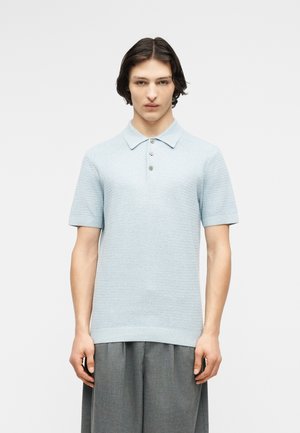 Giovane con capelli castani di media lunghezza che indossa una polo a maglia a maniche corte color azzurro chiaro e pantaloni grigi larghi, in piedi davanti a uno sfondo bianco.