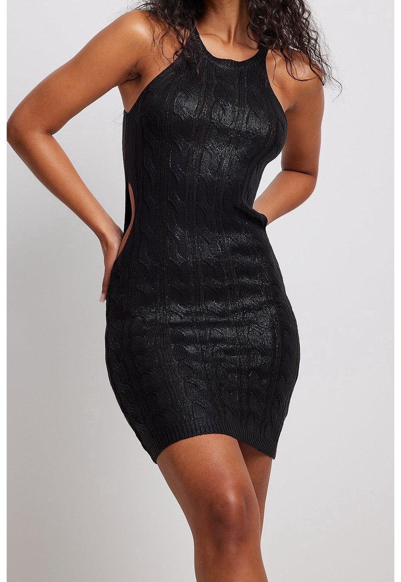 Femme portant une robe mini noire sans manches en maille torsadée avec découpes sur les côtés, debout les mains sur les hanches devant un fond uni.