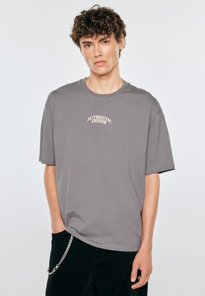 Młody mężczyzna z kręconymi włosami, ubrany w szary t-shirt "Authentic Denim" i czarne spodnie z srebrnym łańcuszkiem, stoi na jednolitym tle.