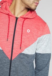 Huvtröja med dragkedja i en tredelad design: röd, grå och mörkgrå. Mjuk textur med röda snörningar och en logotyp på bröstet.