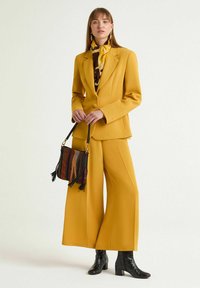 Completo giallo su misura con pantaloni a gamba larga, abbinato a una sciarpa di seta stampata e una borsa multicolore con dettagli a frange e stivali neri.