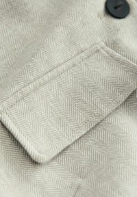 Lichtbeige stof met een visgraatpatroon, met een knoop en een klepzak met zichtbare stiksels. Zachte, gestructureerde oppervlakte.