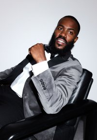 Blazer en velours gris avec revers en satin noir, chemise blanche, nœud papillon noir, assis dans un fauteuil en cuir noir, souriant avec confiance.