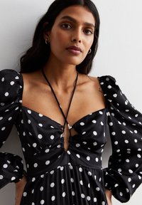Black polka dot dress with a fitted bodice, puff sleeves, and a plunging neckline. Features a pleated skirt and adjustable ties at the back.