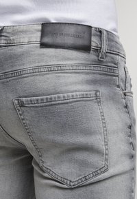 Gros plan sur une personne portant un jean en denim gris clair avec une poche arrière visible et une étiquette en cuir noir sur la ceinture.