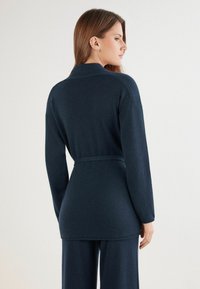 Dunkelblauer Strickcardigan mit langen Ärmeln und einem Bindegürtel in der Taille. Weiche Textur und ein Kragen-Design. Die Rückansicht zeigt eine lockere Passform.