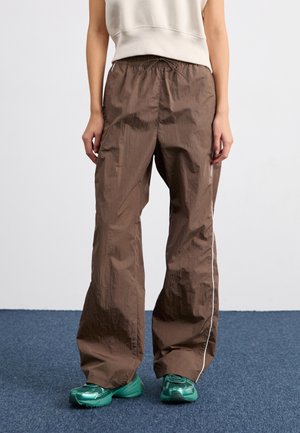 Osoba ubrana w luźne brązowe spodnie z sznurkiem, białe paski po bokach i zielone buty sportowe, stojąca na niebieskim dywanie.