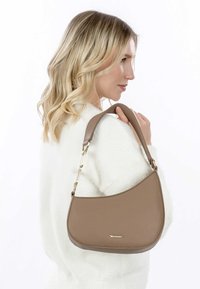 Sac à épaule en simili cuir marron avec une texture lisse, une forme courbée et des accents en métal doré, tenu par une personne aux cheveux blonds.