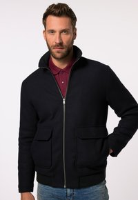 Dunkelblaue Zip-Jacke mit hohem Kragen, zwei Fronttaschen, gerippten Bündchen und glatter Textur, über einem bordeauxfarbenen Poloshirt getragen.