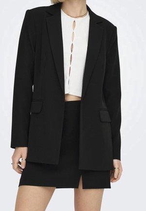 Vrouw draagt een zwarte blazer en rok met een wit cropped top met uitgesneden details aan de voorkant, gecombineerd met ringen en een gouden ketting.