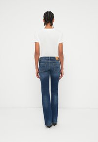 Jean évasés bleu foncé avec une patch en cuir beige sur la ceinture arrière, comportant deux poches arrière et des détails de couture subtils.