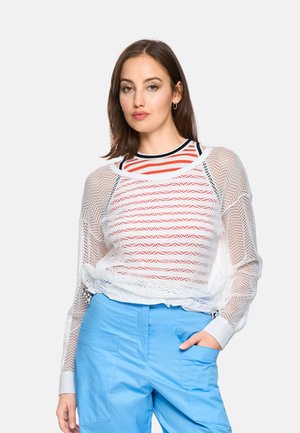 Woman with brown hair wearing a white mesh long-sleeve top over a red and white striped shirt and light blue pants with one hand in pocket.