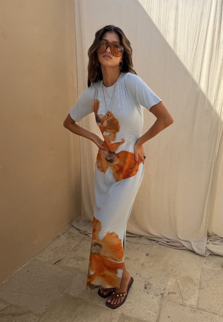 Femme portant une robe maxi blanche avec de grands imprimés fleurs orange, lunettes aviateur, colliers superposés et sandales ornées de perles, posant à l'intérieur.