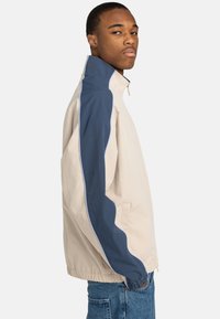 Beige jacket with navy blue accents, stand collar, zip closure, elastic cuffs, and hem, made from lightweight, textured fabric.