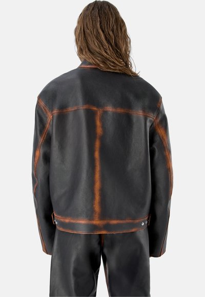 Personne aux cheveux longs portant une veste en cuir noire et un pantalon avec des coutures usées orange-brun, vue de dos sur un fond uni.