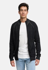 Czarny kardigan na zamek z teksturowanym splotem, z wysokim kołnierzem i ściągaczami na mankietach. Noszony na białej koszuli i jasnoniebieskich dżinsach.