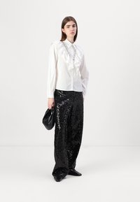 Camisa blanca de botones con detalle de volantes en el frente, combinada con pantalones de pierna ancha negros con lentejuelas. Un bolso de mano negro y unos zapatos planos completan el conjunto.